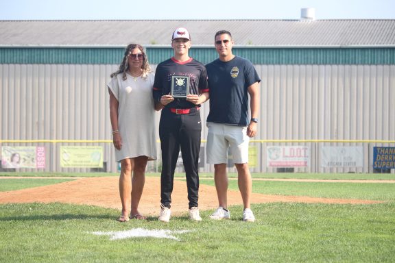 Three people stand on a baseball field, one holding an award, with a fence in the background.